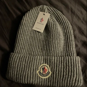 Grå ribbad mössa från Moncler - Säljer en snygg grå ribbad mössa från Moncler. Den är perfekt för kalla dagar och har en klassisk design med Moncler-loggan framtill. Mössan är i nytt skick och passar perfekt till både vardag och vinteraktiviteter.