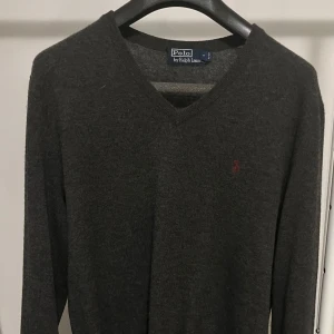 Grå Merino-ulltröja från Polo Ralph Lauren - Säljer en snygg och stilren grå tröja från Polo Ralph Lauren i 100% merinoull. Den har en v-ringad design och det klassiska röda broderade logotypen på bröstet. Perfekt för både höst och vinter, och den är supermjuk och bekväm. Passar perfekt till både vardag och lite finare tillfällen.
