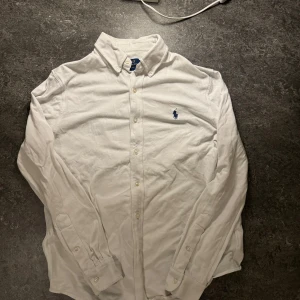 Vit skjorta från Ralph Lauren - Säljer en klassisk vit skjorta från Ralph Lauren i storlek M. Skjortan har långa ärmar och en broderad logga i blått på bröstet. Perfekt för både vardag och fest! 🕺