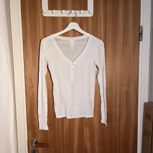 Vit ribbad tröja från H&M - Säljer en stilren vit ribbad tröja från H&M i storlek S. Den har en v-ringad design med knappar framtill och långa ärmar. Perfekt för både vardag och festliga tillfällen. Superbekväm och lätt att matcha med olika outfits! 🤍
