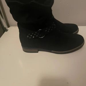 Svarta boots med nitar - Snygga svarta boots i mocka med coola nitar och en dekorativ ringdetalj. Perfekta för höst och vinter, dessa boots har en låg klack och rund tå för bekväm passform. De är i bra skick och redo för nya äventyr!