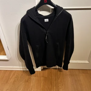 Svart hoodie från C.P. Company - Säljer en stilren svart hoodie från C.P. Company. Den har en dragkedja framtill och praktiska fickor med dragkedja. Perfekt för både höst och vinter, och passar till de flesta outfits. Grymt skön och i bra skick! 