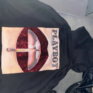Svart hoodie med Playboy-tryck - Säljer en svart hoodie med ett stort, glittrigt Playboy-tryck på ryggen. Perfekt för en cool och avslappnad stil. Hoodien har en klassisk passform med långa ärmar och en bekväm huva. Perfekt för höst och vår!