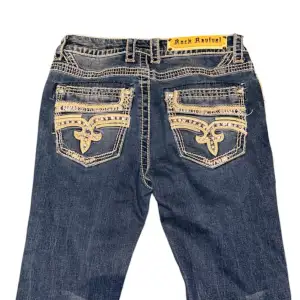 Snygga jeans från Rock Revival i mörkblå denim med unika broderade detaljer på bakfickorna. De har en bootcut-stil och är perfekt för en avslappnad men ändå stilren look. Jeansen har en låg midja och är i bra skick. Perfekta för både vardag och fest! Storlek S, midja är 35cm och innerben är 79cm