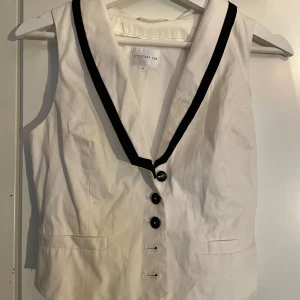Vit väst från Stockh LM - Snygg vit väst från Stockh LM i storlek 38. Den har svarta knappar och en svart kant längs den v-ringade kragen. Västen har en justerbar rem bak för perfekt passform. 