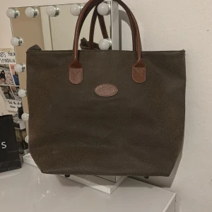 Brun shoppingväska med dragkedja - Säljer en stor och rymlig brun shoppingväska i syntetmaterial. Väskan har två handtag och en praktisk dragkedja upptill. Perfekt för shoppingturer eller som en vardagsväska. Den neutrala färgen gör den lätt att matcha med olika outfits.