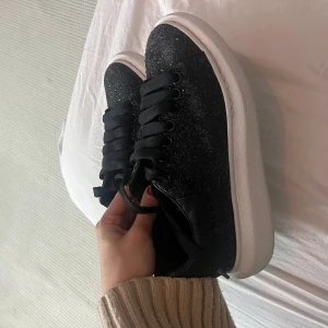 Svarta glittriga sneakers från Alexander McQueen - Säljer ett par svarta glittriga sneakers från Alexander McQueen i mycket bra skick. De har en vit sula och snygg snörning. Perfekta för att ge din outfit lite extra bling! Passar både till vardags och fest. Nypris ligger på 6000 kr