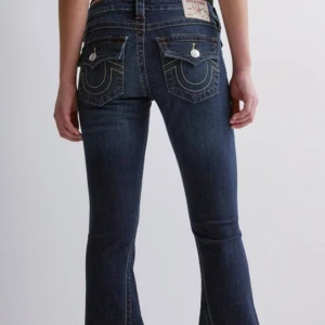 Mörkblå jeans från True Religion - Säljer ett par snygga mörkblå jeans från True Religion. De har en bootcut-stil och är lågmidjade med coola detaljer som knappar och broderade fickor. Perfekta för en avslappnad men stilren look. Passar både till vardags och fest!