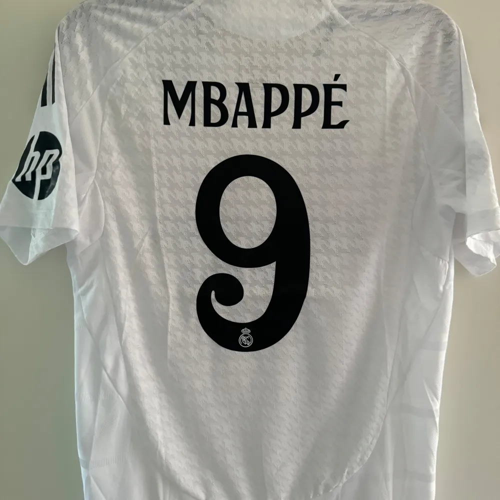 Säljer en vit Real Madrid fotbollströja i storlek L från Adidas. Tröjan har korta ärmar och är i nytt skick. Den har klubbens emblem och sponsorlogga på framsidan samt spelarnamnet 'Mbappé' och nummer 9 på baksidan. Storlek L. T-shirts.