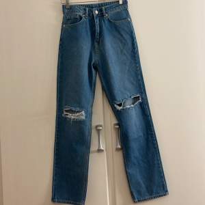 Säljer ett par snygga blå jeans med slitningar på knäna. De har en klassisk femficksdesign och en straight passform. Perfekta för en avslappnad stil och kan matchas med nästan vad som helst. Passar både till vardags och fest!