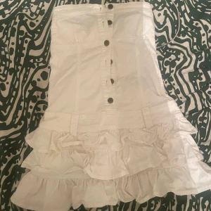 Jeansklänning med volanger - Säljer en superfin vit axelbandslös klänning med volanger. Den har knappar framtill och  volangdetalj längst ner. Klänningen är som ny💕29 rakt över bysten, 32 över midjan och drygt 60 cm lång❤️