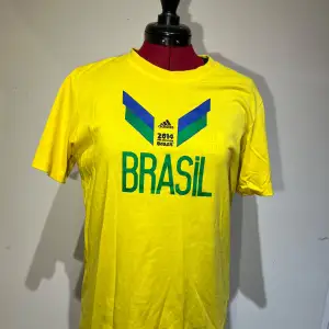 Säljer en gul Brasilien T-shirt från Adidas, officiell produkt från FIFA World Cup 2014. Den har ett tryck med texten 'BRASIL' och färgglada detaljer i blått och grönt. Perfekt för fotbollsfans! 🏆