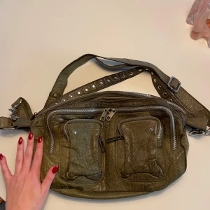 Núnoo axelväska olivgrön  - Säljer en Núnoo axelväska i skinn med nitar på axelremmen. Väskan har flera praktiska fickor med dragkedjor framtill och en justerbar rem. Köparen betalar frakt💖