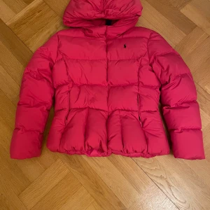 Rosa dunjacka från Ralph Lauren - Säljer en snygg rosa dunjacka från Ralph Lauren i storlek 164 ungefär. Jackan har en mysig huva och är perfekt för kalla dagar. Den är i mycket bra skick och har en klassisk design med quiltade detaljer. Perfekt för både höst och vinter!