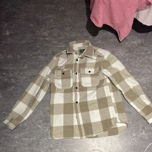Beige och vit rutig skjorta från H&M - Säljer en snygg beige och vit rutig skjorta från H&M. Den har långa ärmar och två bröstfickor med knappar. Perfekt för en avslappnad stil eller lager på lager under hösten. Skjortan är i bra skick och gjord av ett mjukt material som känns skönt mot huden.