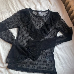 Svart transparent blus med volanger - Snygg svart transparent blus från H&M med prickigt mönster och volangdetaljer. Perfekt för en festlig kväll! Blusen har långa ärmar och en bekväm passform. Passar bra till både jeans och kjol. Använd två gånger