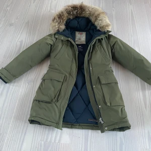 Grön dunparkas från Lexington - Säljer en snygg grön dunparkas från Lexington med pälsfodrad huva. Jackan har dragkedja och knappar framtill samt flera praktiska fickor. Perfekt för kalla vinterdagar! Avtagbar pälskrage