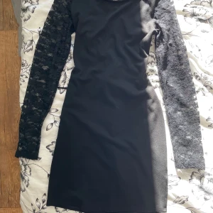 Svart klänning med spetsdetaljer - Snygg svart klänning med långa spetsärmar och en cool snörning i ryggen. Perfekt för en festkväll! Klänningen är tight och har en elegant look med spetsdetaljer som ger en extra touch.