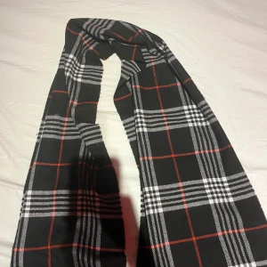 Burrbery halsduk - Säljer en snygg svart halsduk med klassiskt rutigt mönster i vitt och rött. Perfekt för att hålla sig varm under kyliga dagar. Den är stilren och passar till de flesta outfits. Ett måste för höst- och vintergarderoben! Kop 