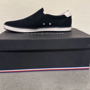 Svarta sneakers från Tommy Hilfiger - Snygga svarta sneakers från Tommy Hilfiger i helt nytt skick aldrig använt . De har en stilren design med vita detaljer och en röd sula. Perfekta för både vardag och fest. Skorna har en bekväm passform och är lätta att matcha med olika outfits.