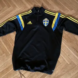 Säljer en svart långärmad fotbollströja från Adidas i Climacool-material. Tröjan har gula och blå detaljer med Sveriges fotbollsförbunds emblem på bröstet. Perfekt för träning eller som supportertröja. Den är i mycket bra skick och redo för nya äventyr på planen! 💙💛