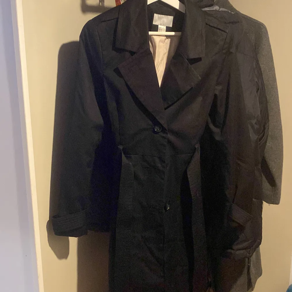 Säljer en stilren svart trenchcoat från H&M i storlek M. Den har en klassisk design med knappar framtill och ett bälte i midjan för en snygg passform. Perfekt för höst och vår! 🖤. Jackor.