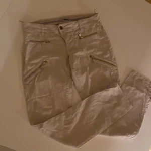 Beige byxor från Hunkydory (Storlek 36) - Säljer ett par snygga beige byxor från Hunkydory. De har en stilren design med dragkedjedetaljer på sidorna och en knapp i midjan. Perfekta för både vardag och fest! (Storlek 36)