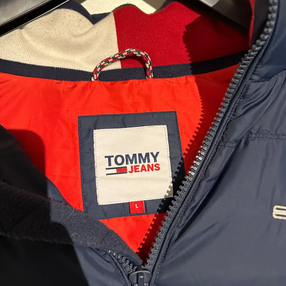 Säljer en snygg mörkblå dunväst från Tommy Jeans i storlek L. Västen har en dragkedja och en cool röd insida som ger en fin kontrast. Perfekt för höst och vinter, håller dig varm och stilren. Passar både till vardags och lite mer avslappnade tillfällen.. Jackor.