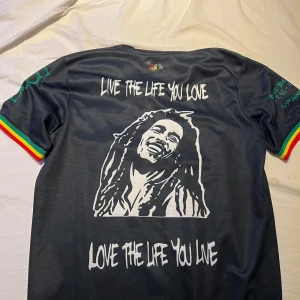 Svart fotbollströja med Bob Marley-tema - Säljer en svart fotbollströja från Adidas med Bob Marley-tema. Tröjan har korta ärmar med rasta-färger på kanterna och flera tryck med citat och bilder av Bob Marley. Perfekt för fotbollsfans som älskar reggae och vill ha en unik stil. Stor logga från Ziggo på framsidan.