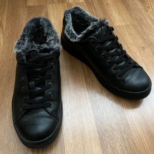 Svarta sneakers med fuskpäls från Calvin Klein - Säljer ett par svarta sneakers från Calvin Klein i mycket bra skick. De har en snygg fuskpälsdetalj runt kanten som gör dem perfekta för höst och vinter. Skorna har snörning och är gjorda i skinn, vilket ger en stilren look. Perfekta för att hålla stilen när det blir kallare!