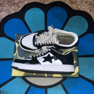 Svartvita sneakers från BAPE - Säljer ett par snygga svartvita sneakers från BAPE i bra skick. De har en glansig finish med den ikoniska stjärndetaljen på sidan. Skorna har snörning och en bekväm sula, perfekt för vardagsbruk eller att sticka ut på stan. Storleken är inte specificerad, men de ser ut att vara i herrstorlek.
