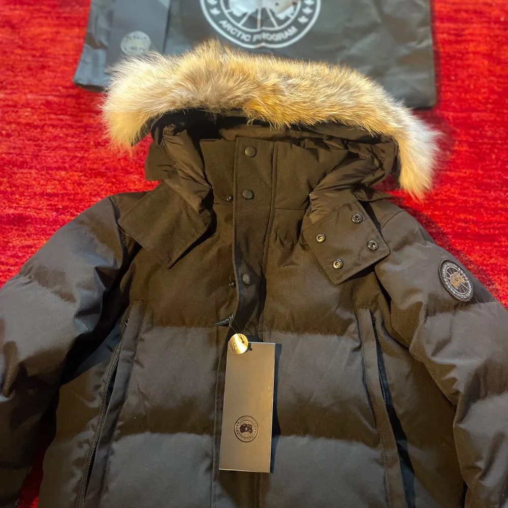 Säljer en svart dunjacka från Canada Goose i mycket bra skick. Jackan har en fluffig pälsdetalj på huvan och både dragkedja och knappar framtill. Perfekt för kalla vinterdagar! Kommer med originalpåse.. Jackor.