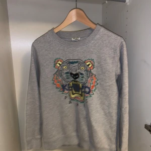  Kenzo - Säljer en grå sweatshirt från Kenzo med ett coolt tigerbroderi på framsidan. Tröjan är i bra skick och perfekt för både höst och vår. Den har långa ärmar och är gjord i mjukt bomullsmaterial. Passar perfekt för en avslappnad stil eller en dag på stan.