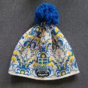 Mönstrad mössa från Craft - Säljer en superfin mössa från Craft med ett unikt mönster i blått, gult och vitt. Den har en stor blå tofs på toppen och är perfekt för vintern. Mössan är gjord i polyester och ull, vilket gör den både varm och bekväm. Perfekt för skidåkning eller kalla dagar! 🇸🇪