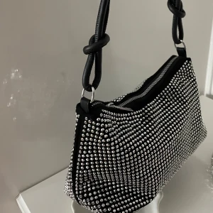 Svart axelväska med silverdetaljer - Trendig svart axelväska täckt med glittrande silverpärlor. Väskan har en dragkedja upptill och en stilren design med knutna axelremmar. Använd bara en gång✨✨
