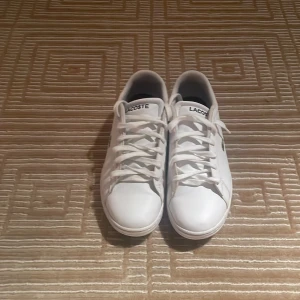 Vita sneakers från Lacoste - Snygga vita sneakers från Lacoste med klassisk design och snörning. Skorna har den ikoniska krokodilloggan på sidan och är perfekta för en stilren look.