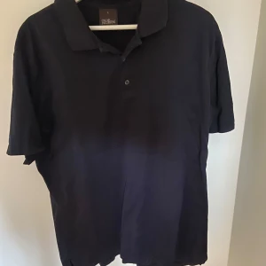 Svart pikétröja från Oscar Jacobson - Snygg svart pikétröja från Oscar Jacobson i storlek L. Perfekt för en stilren och avslappnad look. Tröjan har korta ärmar och en klassisk krage med knappar framtill.