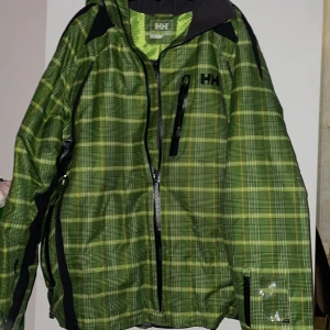 Ny Helly Hansen Grön rutig jacka   - Säljer en grön rutig jacka från Helly Hansen ny med dragkedja och flera praktiska fickor. Jackan har en sportig look med långärmade ärmar och är perfekt för utomhusaktiviteter. Den har ett meshfoder på insidan för extra komfort.