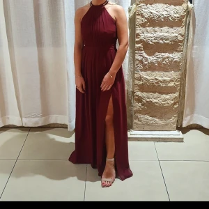 Vinröd långklänning med öppen rygg - Elegant vinröd långklänning med halterneck och öppen rygg. Klänningen har en hög slits och dekorativa detaljer vid halsen. Perfekt för festliga tillfällen.