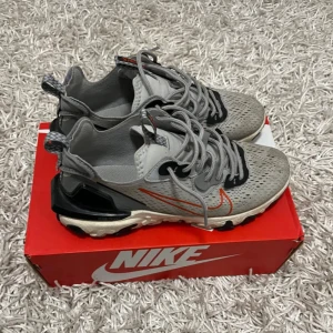 Nike React Vision  - Snygga grå Nike sneakers med orange swoosh och svarta detaljer. Skorna har en modern design med snörning och perforerat mönster för extra ventilation. Perfekta för en sportig look.