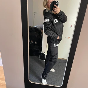 Svart hoodie med dragkedja och tryck - Snygg svart hoodie med dragkedja och vita tryckdetaljer på bröstet och ärmen. Perfekt för en avslappnad stil. Hoodien har en bekväm passform och är idealisk för vardagsbruk.Fudrade.Helt ny