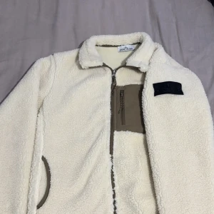 Vit fleecejacka från Stone Island - Säljer en snygg vit fleecejacka från Stone Island med dragkedja och en broderad logotyp på ärmen. Jackan har en praktisk bröstficka med dragkedja och är perfekt för kyliga dagar.