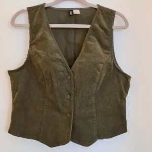 Olivgrön manchester väst från H&M - Snygg olivgrön väst i manchester från H&M. Väst med knappar framtill och en v-ringad design. Perfekt för att lägga till en touch av stil till din outfit.