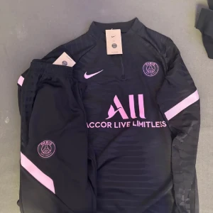 Svart och rosa fotbollströja från Nike - Snygg svart fotbollströja från Nike med rosa detaljer och PSG-logga. Tröjan har en halv dragkedja och långa ärmar. Perfekt för fotbollsfans som vill ha stil på planen.