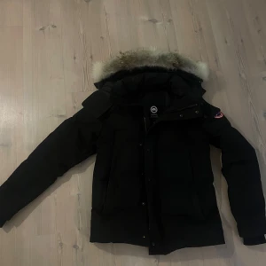 Svart Canada goose wyndham - Säljer en svart parkajacka från Canada Goose med pälskantad huva. Jackan har långa ärmar och dragkedja framtill. Perfekt för kalla vinterdagar. Säljer jackan pga att jag har tröttnat på den. 