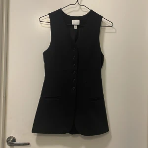 Svart lång väst med knappar - Snygg svart väst med knappar framtill. Perfekt för en stilren look. Västen har en v-ringad design och är figursydd för en elegant passform.