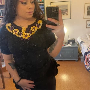 Svart topp med broderade detaljer - Snygg svart topp med korta ärmar och färgglada broderade detaljer i gult och lila runt halsringningen. Perfekt för att ge en unik touch till din outfit.