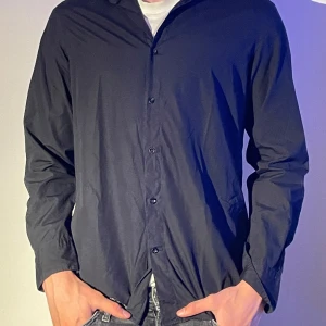 Svart skjorta från Massimo Dutti - Elegant svart skjorta från Massimo Dutti i ett fint tyg. Skjortan har långa ärmar och knappar framtill, perfekt för en stilren look. Tillverkad med högkvalitativt material.