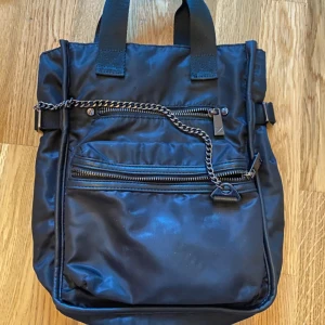 Svart ryggsäck från Zara - Snygg svart ryggsäck från Zara i syntetmaterial. Den har flera dragkedjor och en kedjedetalj på framsidan. Perfekt för en stilren look.