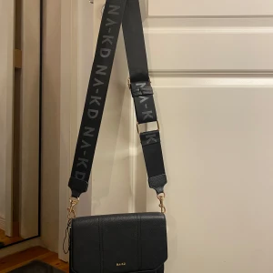 Svart axelväska från NA-KD - Snygg svart axelväska från NA-KD med justerbar rem och gulddetaljer. Väskan har en texturerad yta och insida med ormmönster. Perfekt för att bära dina nödvändigheter med stil.